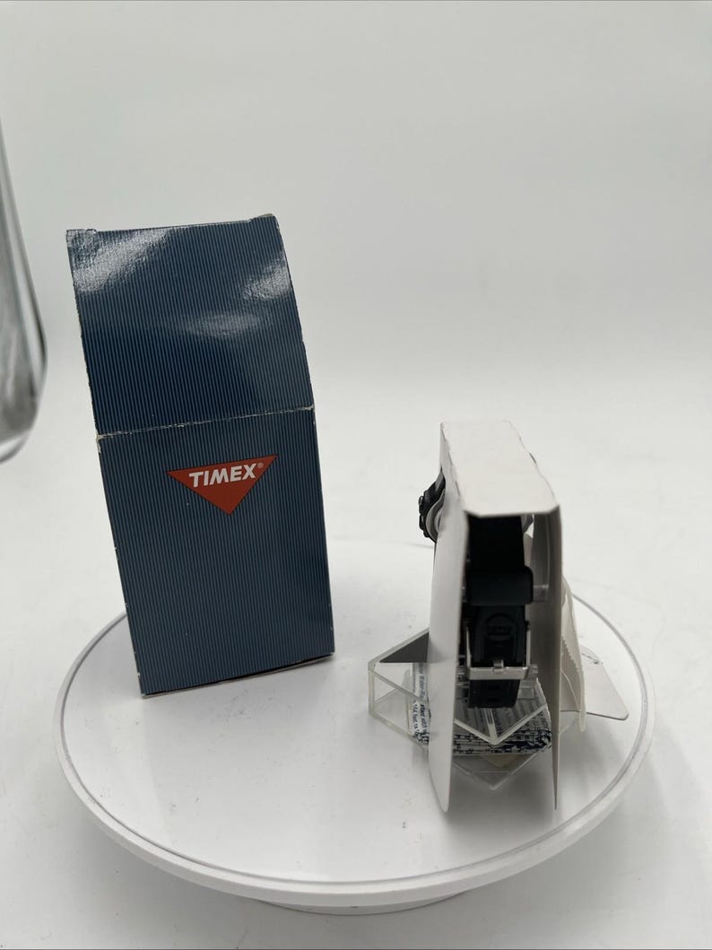 May include: A black Timex watch with a black band is displayed on a white surface. The watch is presented in its original packaging, alongside a navy blue box with the Timex logo.