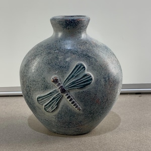 May include: A round, gray ceramic vase with a textured surface and a dragonfly design. The dragonfly is teal with a brown body. The vase has a narrow neck and a wide, rounded body.