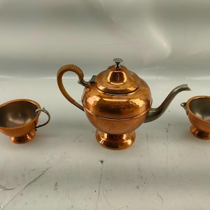 May include: A copper tea set with a teapot, creamer, and sugar bowl. The teapot has a woven handle and a silver spout. The creamer and sugar bowl have small handles. The set is made of copper with a hammered texture.