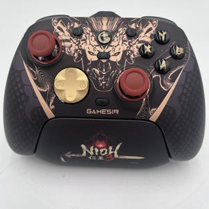 Puede incluir: Un mando de videojuegos GameSir negro con un diseño gráfico de Nioh 3 en cobre y negro. El mando incluye una cruceta dorada, dos joysticks rojos y botones de acción negros con símbolos dorados sobre una superficie oscura texturizada.