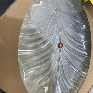 May include: Oval-shaped clear glass serving platter with a frosted floral design in the center. The platter has a textured, rippled pattern radiating from the flower. A small, round sticker is visible near the bottom.