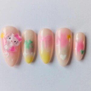 Peut inclure: Ensemble de cinq faux ongles avec une base rose clair. Les motifs comprennent Hello Kitty, un trèfle à quatre feuilles et des dégradés roses et jaunes avec des cœurs et des soleils. Les ongles sont de forme ovale.