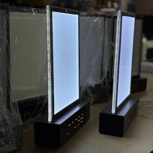Könnte beinhalten: Mehrere beleuchtete LED-Displays mit rechteckiger Form und schwarzem Sockel. Die Schilder haben ein helles weißes Licht und das Wort "COSE" ist auf dem Sockel sichtbar. Die Schilder stehen auf einer hellgrauen Oberfläche.