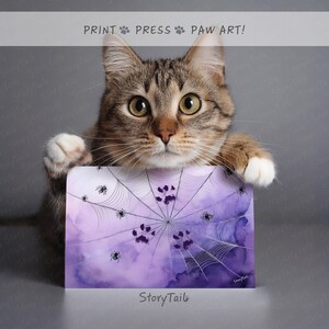 May include: A tabby cat holds a rectangular print featuring a watercolor spiderweb design in shades of purple. Black spiders and paw prints are scattered across the web. The print has the text "PRINT PRESS PAW ART!" at the top and "StoryTail" at the bottom.