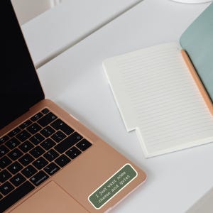Peut inclure: Un ordinateur portable rose dor&eacute; avec un clavier noir et un autocollant vert qui dit "I just want some cheese and quiet." Un cahier blanc et un crayon sont &eacute;galement visibles sur le bureau blanc.
