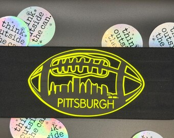 Puede incluir: Artículo rectangular negro con un diseño de fútbol amarillo neón, con un horizonte y la palabra "PITTSBURGH". Alrededor hay pegatinas circulares con el texto "think outside the can".
