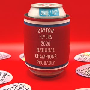 May include: A red can cooler with white, blue, and red trim, holding a can. The cooler reads "DAYTON FLYERS 2020 NATIONAL CHAMPIONS PROBABLY." The can reads "0 Sugar Carbs Gluten Free." Scattered around are stickers that read "think outside the can."