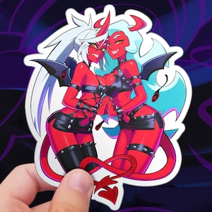 May include: A sticker featuring two red-skinned anime-style characters with horns, wings, and devil tails. They wear black leather outfits and have white and light blue hair. The sticker is held by a hand.