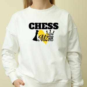 May include: White sweatshirt with the text "CHESS Mom" in black, with a yellow heart shape. The design includes a chess piece and a crown. The sweatshirt is a casual style.