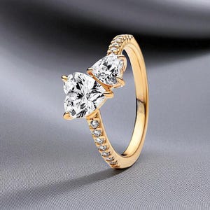 Puede incluir: Un anillo dorado con dos gemas claras en forma de corazón. La banda también está adornada con pequeñas gemas claras. El anillo está sobre un fondo gris.