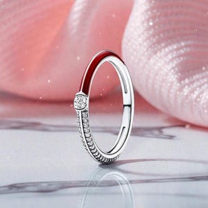 Peut inclure: Une bague en argent avec un accent en émail rouge et une rangée de petites pierres claires. Une seule pierre claire plus grande est sertie en haut de la bague. La bague est présentée sur une surface réfléchissante avec un fond rose.