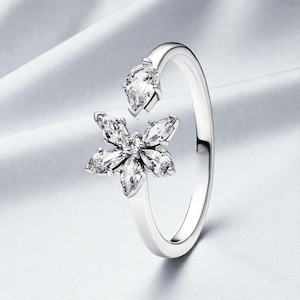 Puede incluir: Anillo plateado con diseño floral. El anillo presenta piedras preciosas claras y brillantes dispuestas en forma de flor, con una piedra en forma de lágrima en la parte superior. El fondo blanco resalta su elegante diseño.