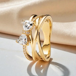 以下が含まれることがあります： ゴールドカラーのリングセット。1つのリングは、小さなクリアストーンと2つの大きな三角形のクリアストーンで飾られたバンドが付いています。もう1つのリングは、滑らかで絡み合ったデザインです。中立的な背景。