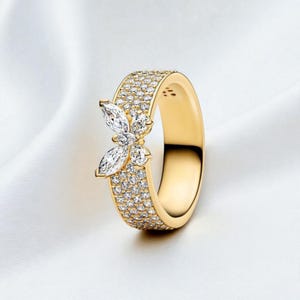 Puede incluir: Un anillo de oro con un diseño de mariposa. La banda está incrustada con pequeñas piedras brillantes. La mariposa está hecha de piedras más grandes, con corte marquesa. El anillo se presenta sobre un fondo blanco.