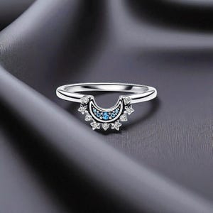 Puede incluir: Un anillo plateado con un diseño en forma de media luna. El anillo presenta una fila de pequeñas piedras azul claro en el centro de la media luna, rodeadas de acentos claros en forma de estrella. El anillo está sobre un fondo gris oscuro.