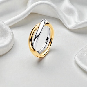 Pode incluir: Um anel de duas cores com uma banda dourada e uma banda prateada que se cruzam. O anel é apresentado sobre um fundo branco.