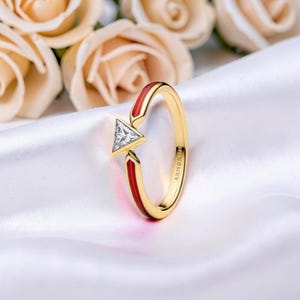 May include: A gold ring with a triangular, clear gemstone. The ring has red enamel accents and the word "ARMOR" is engraved on the band. The ring is set against a white satin background with soft focus roses.