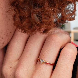 May include: A gold ring with a red enamel band and a small, triangular, clear gemstone. The ring is worn on a finger, with the focus on the jewelry's design and details. The ring's design is simple and elegant.