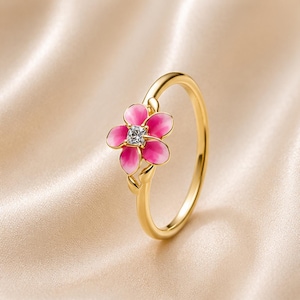 Puede incluir: Un anillo de oro con un motivo floral de cinco pétalos con detalles en esmalte rosa degradado y una piedra central transparente de corte cuadrado. La banda es de oro liso y pulido, con pequeños acentos en forma de hoja en la base de la flor.