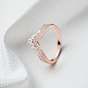 Könnte beinhalten: Roségoldfarbener Ring mit V-förmigem Design, verziert mit funkelnden, klaren Edelsteinen. Der Ring hat ein zartes Band und eine kronenartige Spitze, vor einem weißen, strukturierten Hintergrund. Ein Schmuckstück.