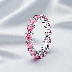 Puede incluir: Un anillo de plata con una fila continua de piedras preciosas rosas en forma de corazón. El anillo está engastado sobre un fondo blanco texturizado, resaltando el brillo de las piedras preciosas. El diseño es elegante y romántico.