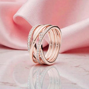May include: A rose gold ring featuring multiple interwoven bands, some adorned with small, clear gemstones. The ring is displayed against a soft pink backdrop, giving a luxurious and elegant feel. This is a piece of jewellery.