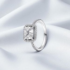 Peut inclure: Une bague en argent ornée d'une grande pierre précieuse rectangulaire et transparente, entourée d'un halo de petites pierres transparentes. Le corps de la bague est également orné de petites pierres transparentes. La bague est posée sur un fond blanc texturé.