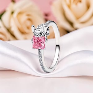K&ouml;nnte beinhalten: Ein silberner Ring mit einer Cartoonfigur, die einen quadratischen, rosa Edelstein h&auml;lt. Das Ringband ist mit kleinen, klaren Edelsteinen verziert. Der Ring ist vor einem weichen, wei&szlig;en Stoffhintergrund mit verschwommenen rosa Blumen platziert.