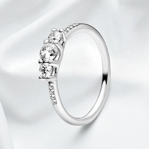 Könnte beinhalten: Ein silberner Ring mit drei großen, klaren Edelsteinen in einer Reihe. Das Band ist ebenfalls mit kleineren, klaren Edelsteinen besetzt. Der Ring wird auf einer weißen, strukturierten Oberfläche präsentiert, die sein elegantes Design und seinen Glanz hervorhebt.