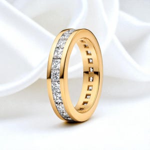 Puede incluir: Un anillo dorado con una banda de piedras preciosas cuadradas y transparentes. El anillo se muestra sobre un fondo blanco y sedoso, resaltando su diseño y brillo. El anillo es una pieza de joyería.