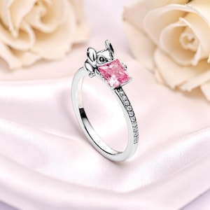 K&ouml;nnte beinhalten: Silberner Ring mit einer Cartoonfigur, die einen quadratischen, rosa Edelstein h&auml;lt. Das Ringband ist mit kleinen, klaren Edelsteinen verziert. Der Ring befindet sich vor einem weichen, verschwommenen Hintergrund.