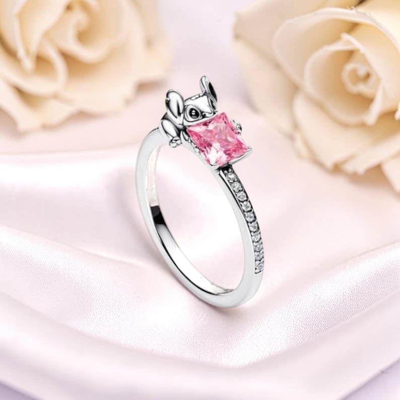 K&ouml;nnte beinhalten: Silberner Ring mit einer Cartoonfigur, die einen quadratischen, rosa Edelstein h&auml;lt. Das Ringband ist mit kleinen, klaren Edelsteinen verziert. Der Ring befindet sich vor einem weichen, verschwommenen Hintergrund.