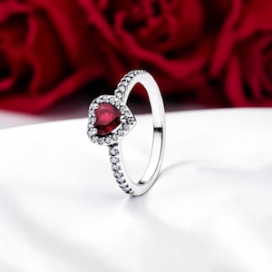 Puede incluir: Un anillo plateado con una piedra preciosa roja oscura en forma de corazón, rodeada de pequeñas piedras transparentes. La banda también está adornada con pequeñas piedras transparentes. El anillo está sobre un fondo blanco con un fondo floral rojo borroso.