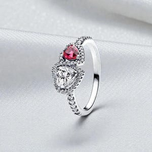 Peut inclure: Une bague en argent ornée de deux pierres en forme de cœur. L'une est un cristal clair et facetté, l'autre une pierre rouge vif. L'anneau est orné de petits accents scintillants, créant un design délicat et élégant.