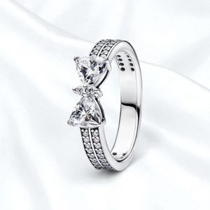 Puede incluir: Un anillo plateado con un diseño en forma de lazo. El anillo presenta dos piedras claras en forma de corazón y piedras claras más pequeñas engastadas a lo largo de la banda. El anillo está sobre un fondo blanco.