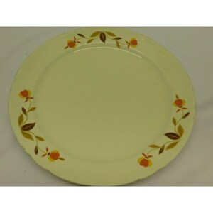 May include: A round, cream-colored plate with a scalloped edge. The plate is decorated with a floral pattern featuring orange and yellow flowers with brown leaves. The design is arranged around the rim of the plate.
