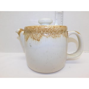 May include: A ceramic teapot with a lid, featuring a white base and a brown, textured rim. The teapot has a curved handle and a spout for pouring. The lid has a small, round knob. A ruler is visible in the background.