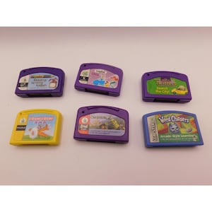 Pode incluir: Seis cartuchos de jogos educativos LeapPad em várias cores. Os títulos incluem "Reading, Writing & Math", "LeapPad", "Twisters Search the City!", "Vroom Vroom! On the Go", "Shrek 2" e "Word Chasers". Os cartuchos são projetados para a aprendizagem infantil.
