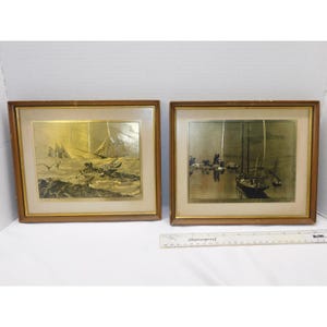 Op de afbeelding: Twee ingelijste nautische kunstprints. De prints tonen goudkleurige afbeeldingen van zeilboten op het water. De frames zijn middelbruin met een gouden binnenrand. Eén print toont zeilboten in woelig water, de andere boten in een haven.