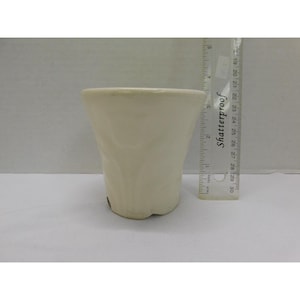 May include: A cream-colored ceramic cup with a textured exterior. The cup has a wide opening and a slightly flared shape. A clear ruler with the word "Shatterproof" is next to the cup.