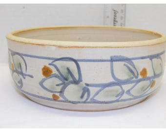 May include: A ceramic bowl with a cream-colored exterior and a blue and green floral pattern. The bowl has a wide, shallow shape and a light brown rim. The floral design is outlined in blue and features green leaves and brown accents.