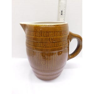 May include: A brown ceramic pitcher with a handle and a spout. The pitcher has a barrel design with vertical wood grain and horizontal bands. The interior is white. A ruler is in the background.