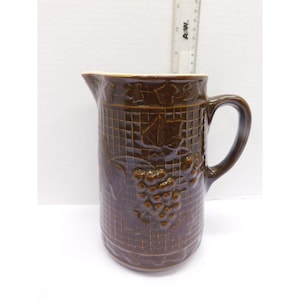 May include: Brown ceramic pitcher with a handle and spout. The pitcher features a grid pattern and embossed grape and leaf design. The pitcher is approximately 20 cm tall.