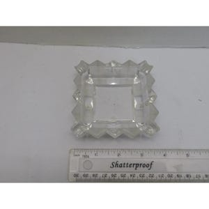 Op de afbeelding: Transparante, vierkante asbak met een geometrisch, facetachtig ontwerp. De asbak heeft een centrale vierkante verdieping en vier hoeksteunen. Een liniaal met de tekst "Shatterproof" staat in de afbeelding.
