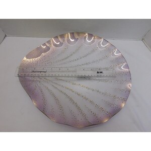 May include: A decorative, shell-shaped plate with a wavy rim. The plate is primarily white with a subtle lavender hue and gold speckles. A clear ruler with the text "Shatterproof" is placed across the plate. The plate measures approximately 12 inches in diameter.