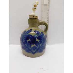 Pode incluir: Jarro de cerâmica com uma pega, apresentando desenhos florais azuis sobre um fundo bege. Possui uma rolha de cortiça e um bico decorativo. O jarro tem aproximadamente 18 cm de altura.