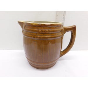 May include: A brown ceramic pitcher with a barrel-like design. The pitcher has a handle and a spout for pouring. The exterior features horizontal bands and vertical lines, giving it a rustic appearance. The pitcher is likely used for serving beverages.