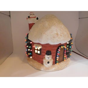 May include: A ceramic holiday house figurine featuring a red brick exterior, a snow-covered roof, and a chimney with Santa Claus emerging from the top. A small snowman stands by the brown door, and the structure is decorated with colorful lights.