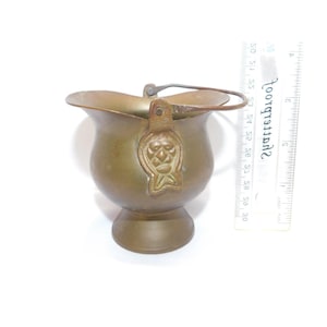 May include: A small, antique brass bucket with a handle. The bucket has a decorative emblem on the side and sits on a pedestal base. The bucket's opening is approximately 3 inches wide.