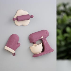 Op de afbeelding: Een verzameling keukenmagneten in paars en crème. De set bevat een deegroller op deeg, een ovenwant en een keukenmixer met een kom, perfect voor het decoreren van een koelkast of ander metalen oppervlak.